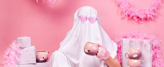 A ghost in a white sheet wearing pink heart-shaped sunglasses holds a pink Cloud mug printed with a bat, beside stacked Egg Back Home gift boxes and pink feather boas